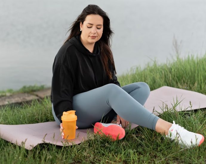 Woman enjoying a peaceful moment by the lake with a yoga mat and water bottle, promoting wellness.