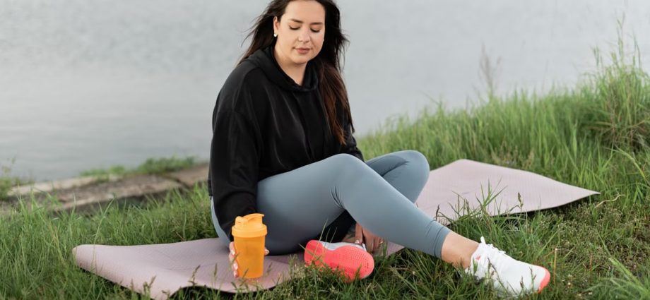 Woman enjoying a peaceful moment by the lake with a yoga mat and water bottle, promoting wellness.
